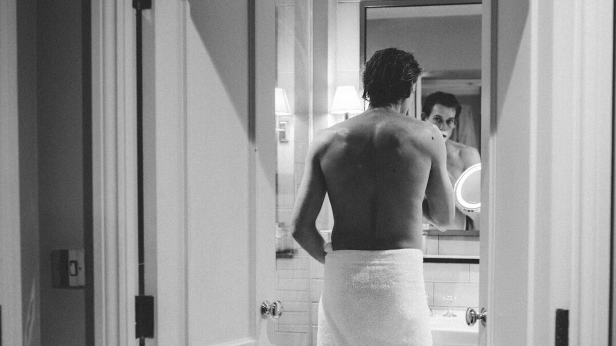 Attractive man applying a safety razor to his foam-covered face while shaving in front of a bathroom mirror during a traditional wet shave